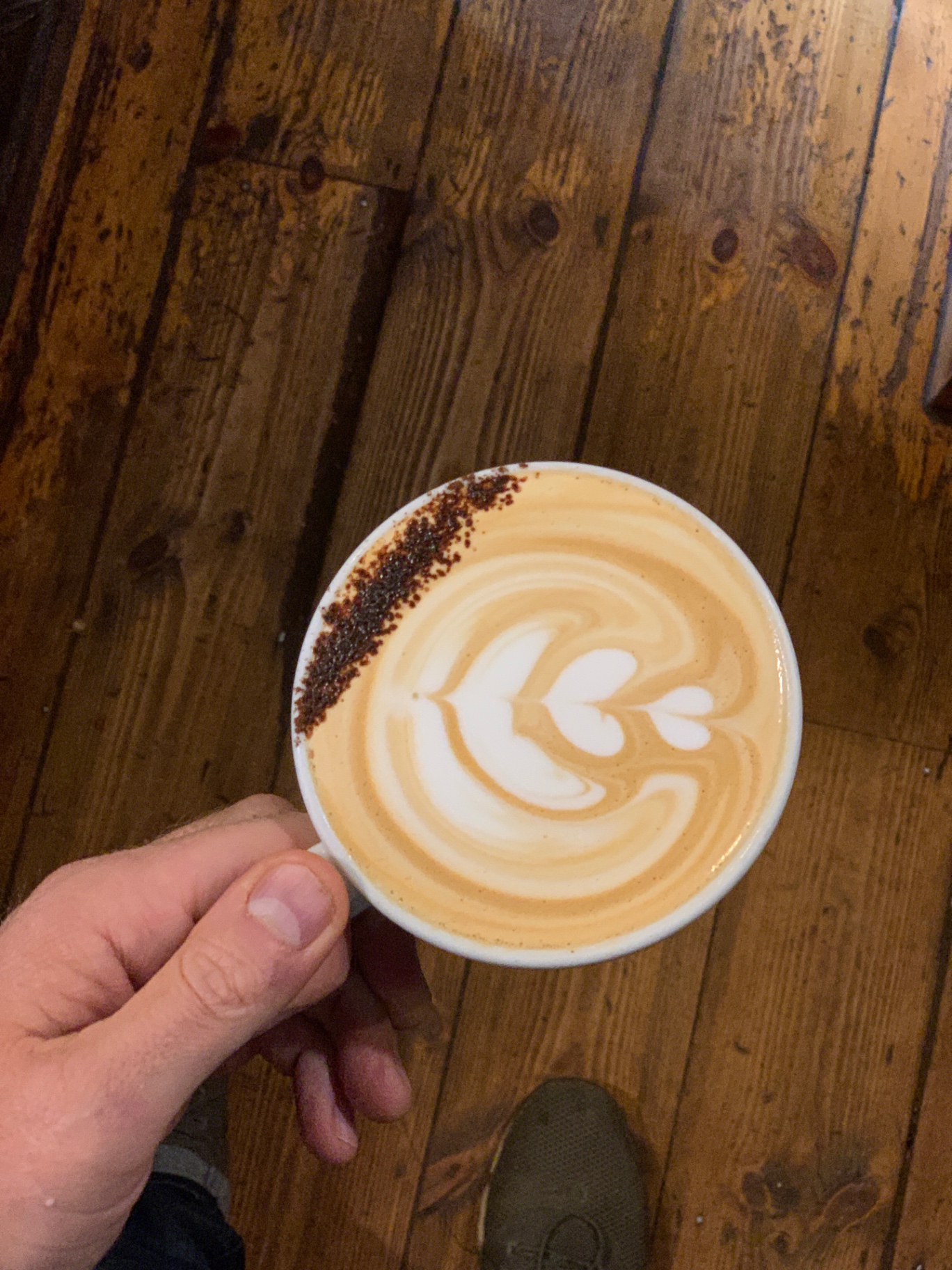 Flat white with tulip latte art and a stripe of dusted cocoa, held over rustic wooden floorboards
